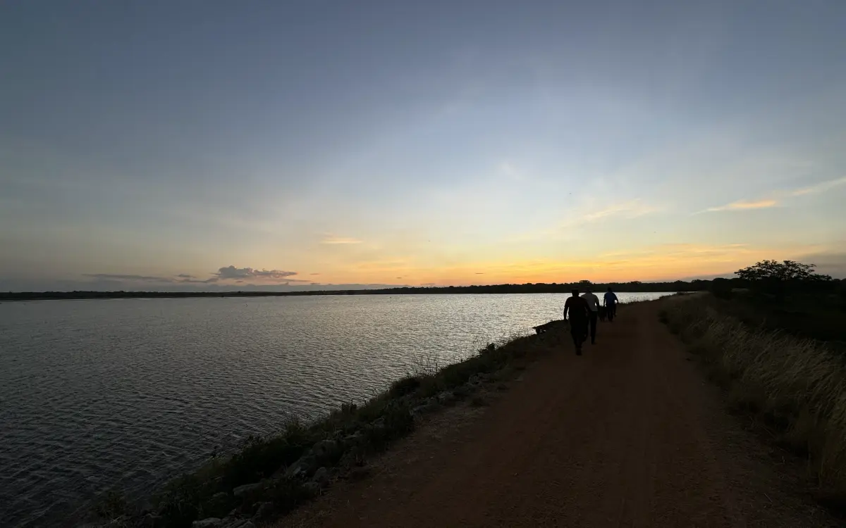 People walking the earthen bund of a tank at sunset, water on one side